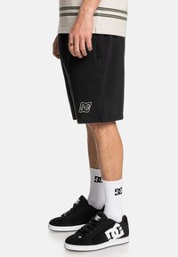 Zwarte katoenen shorts met zijzakken en een wit DC-logo. Gecombineerd met zwarte sneakers en witte sokken met een logo ontwerp.