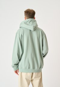 Hooded sweatshirt in light mint green, made from soft material. Features a relaxed fit with ribbed cuffs and hem. Wall visible in white background.