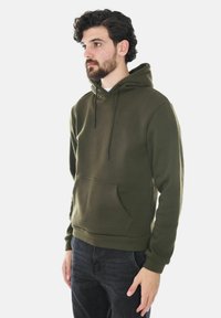 Felpa verde oliva, realizzata in tessuto morbido, presenta una tasca frontale, coulisse e polsini a coste. Il modello indossa jeans scuri.