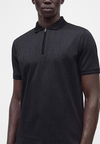 Homme portant un polo noir à manches courtes avec une texture subtilement travaillée et une fermeture éclair frontale au lieu de boutons.