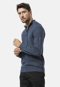 Pull en maille bleu marine à fermeture éclair, avec des poignets et un ourlet côtelés. Détail de logo sur le côté du bras gauche.