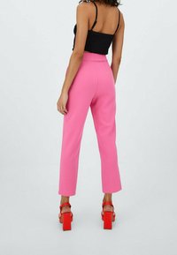 Pantalon rose sur mesure avec une coupe droite, une taille mi-hauteur et une texture lisse. Porté avec des sandales à talons épais rouge vif.