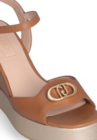 Brown leather wedge sandal with an open toe, gold hardware logo detail, and textured light beige platform sole.