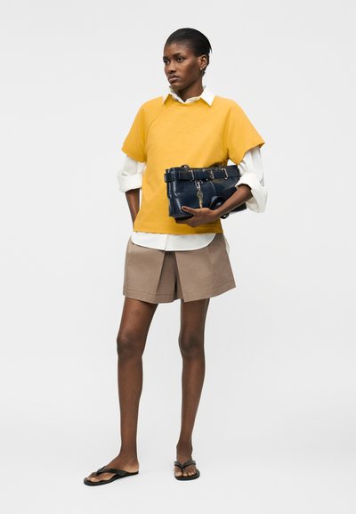 Femme portant un sweat-shirt jaune à manches courtes par-dessus une chemise blanche, un short beige, des tongs noires, tenant un sac à main bleu marine avec un porte-clés en forme de clé.