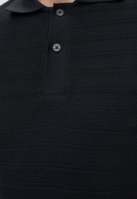 Gros plan d’un polo en maille noire texturée avec un col et deux boutons noirs fermés au niveau de l’encolure.