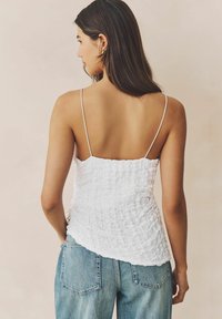 Camisola bianca con texture e spalline sottili, abbinata a jeans a vita alta di colore blu chiaro. La parte superiore ha una silhouette arricciata e un orlo irregolare.