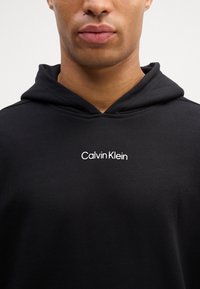 Črna kapuca iz mešanice bombaža; ima majhen bel "Calvin Klein" logotip na prsih, gladko teksturo in sproščen kroj.