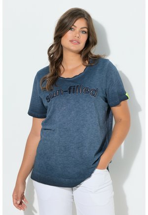 Vrouw met lang bruin haar, een blauw "zonovergoten" T-shirt en witte broek, staand met één hand in de zak tegen een witte achtergrond.