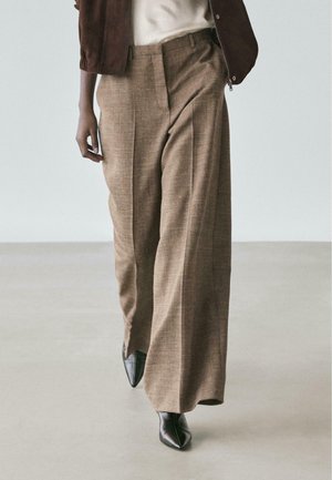 Personne portant un pantalon marron taille haute à jambes larges, des chaussures pointues noires, un haut blanc et une veste marron foncé marchant sur un sol clair.