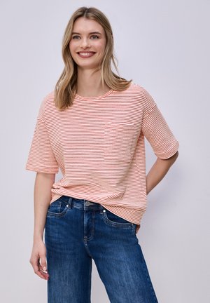 Junge Frau lächelt, trägt ein kurzärmeliges, hellrosa-rot gestreiftes Shirt mit Brusttasche und blaue Jeans, steht vor einem schlichten Hintergrund.