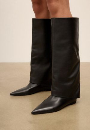 Bottes en cuir noir longues jusqu'au genou avec des bouts pointus et des semelles plates portées sur des jambes nues sur un fond beige uni.