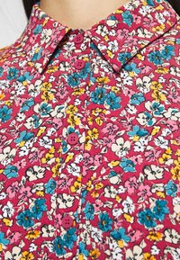 Camicia button-up floreale con una base rosa, piccoli fiori multicolori e bottoni rosa. Design con colletto e texture fine.