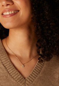 Gouden ketting met een bloem pendant met heldere edelstenen, gedragen boven een gestructureerde, beige V-hals trui.