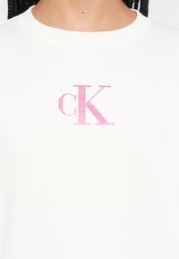 Biela mikina s ružovým pixelovaným logom "CK" na hrudi. Materiál pôsobí hladko a má klasický okrúhly výstrih.