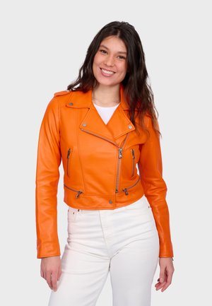 Jonge vrouw die glimlacht, gekleed in een oranje leren jas over een witte top en witte broek, staand tegen een effen lichte achtergrond.