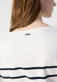 Witte gebreide top met marineblauwe horizontale strepen en een zilveren logo label aan de achterkant van de halslijn. Zachte textuur en ontspannen pasvorm ontwerp.