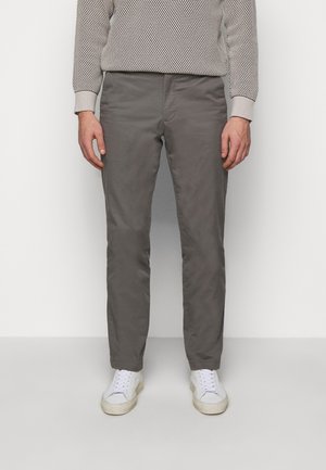 Uomo che indossa pantaloni grigi dritti, scarpe da ginnastica bianche e un maglione a maniche lunghe strutturato, in piedi contro uno sfondo bianco neutro.