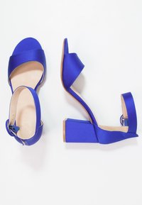 Pár otevřených sandálů na blokovém podpatku v královsky modré saténové barvě s kotníkovými pásky a stříbrnými přezkami na bílém pozadí.