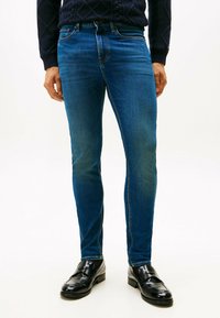 Uomo che indossa jeans slim-fit blu scuro, mocassini in pelle nera e un maglione strutturato blu navy, in piedi contro uno sfondo bianco uniforme.
