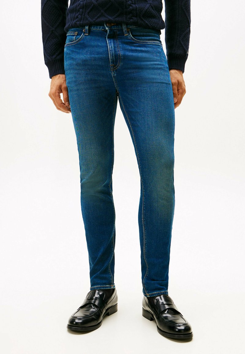 Uomo che indossa jeans slim-fit blu scuro, mocassini in pelle nera e un maglione strutturato blu navy, in piedi contro uno sfondo bianco uniforme.