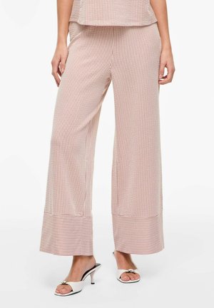 Nő bézs és fehér csíkos, bőszárú nadrágot és hozzáillő felsőt visel, fehér, nyitott orrú, masnis magassarkú szandállal párosítva.