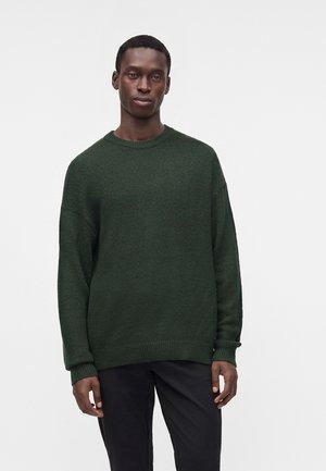 Mand iført en mørkegrøn strikket langærmet sweater og sorte bukser, stående foran en ensfarvet lys baggrund.
