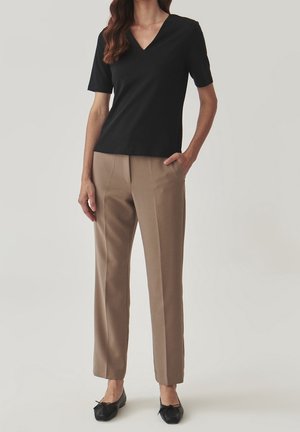 Woman wearing black short-sleeve V-neck top, beige straight-leg trousers, and black ballet flats, standing with one hand in pocket.