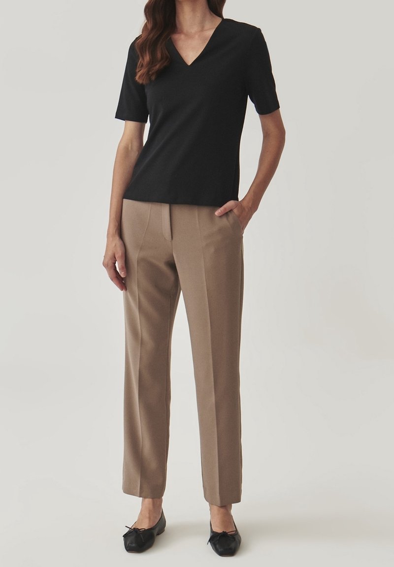 Femme portant un haut noir à manches courtes et col en V, un pantalon beige à jambes droites et des ballerines noires, debout avec une main dans la poche.