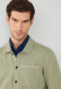Giacca verde oliva chiaro con tasca frontale, chiusura a bottoni e colletto strutturato. Indossata su una camicia blu scuro.