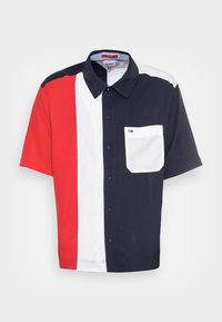 Košile s krátkým rukávem, která má vertikální bloky v červené, bílé a námořnické barvě, a bílou náprsní kapsu s malým logem.
