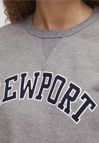 Felpa grigia con colletto a coste, caratterizzata da una grafica curva "NEWPORT" in blu navy. Il tessuto presenta una consistenza morbida con un leggero lucido.