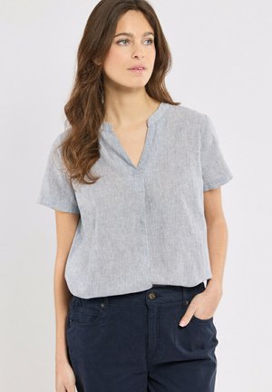 Femme aux longs cheveux bruns portant un chemisier à manches courtes gris clair et un pantalon bleu marine foncé, une main dans la poche, regardant sur le côté.