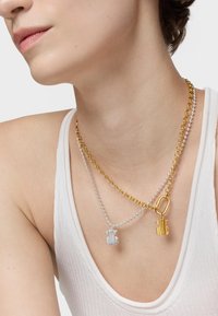 Deux colliers empilés : une chaîne dorée avec un charme en forme d'ours, une chaîne argentée à billes avec un charme d'ours rayé. Fond et haut neutres.