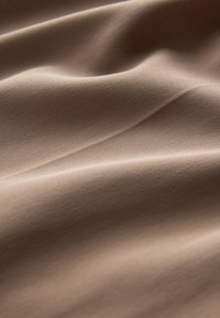 Pehmeä, sileä beige kangas, jossa hienovarainen vaakasuora ribbineulos ja lempeät laskokset luovat varjoja ja tekstuuria pinnalle.