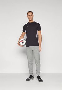 Puma melna īsām piedurknēm krekls ar pelēkiem sānu akcentiem, pelēkas sporta bikses un melni apavi. Modelis tur rokās futbolbumbu.