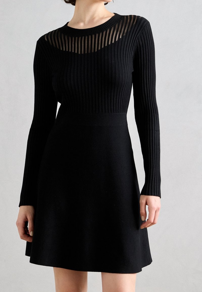 Femme portant une robe noire en maille côtelée à manches longues avec un empiècement transparent à rayures et une jupe évasée, debout devant un fond uni.