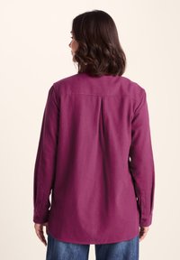 Long-sleeve shirt in deep magenta, structured fabric with a relaxed fit, featuring buttoned cuffs and a yoke seam at the back.