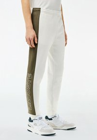 Bílé teplákové kalhoty s tmavě zeleným bočním panelem a vytištěným logem "LACOSTE". Disponují elastickým páskem a zúženým střihem, doplněné bílými teniskami.