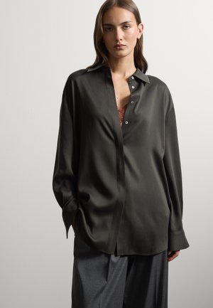 Vrouw draagt losse zwarte satijnen blouse met knopen over een kanten top en grijze wijde broek tegen een effen lichte achtergrond.