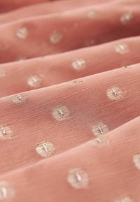 Tissu rose doux avec un tissage texturé subtil et de petits points carrés dorés brodés et brillants, uniformément répartis sur la surface.