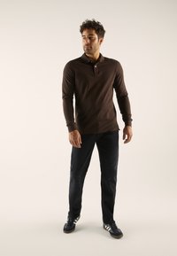 Moški stoji in nosi temno rjavo polo majico z dolgimi rokavi, črne kavbojke ter črne superge z belimi črtami na enobarvni podlagi.