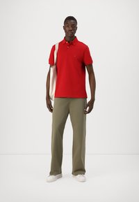 Camisa polo roja, de manga corta, con cuello; pantalones rectos de color verde oliva; zapatillas blancas; bolsa de lona; diseño minimalista, textura lisa.