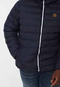 Man in een donkerblauwe gewatteerde jas met een rits aan de voorkant en een blauwe spijkerbroek, met de rechterhand in de zak en de linkerhand ontspannen naast het lichaam.