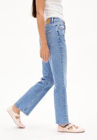 Jean en denim bleu clair avec une coupe droite, une finition délavée, cinq poches et un patch en cuir marron à la taille arrière. Associé à des ballerines roses.