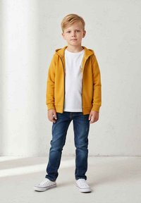 Żółta bluza na zamek z kapturem nałożona na białą koszulkę, zestawiona z niebieskimi dżinsami i białymi trampkami. Tkanina wydaje się miękka; dżinsy są lekko postrzępione.