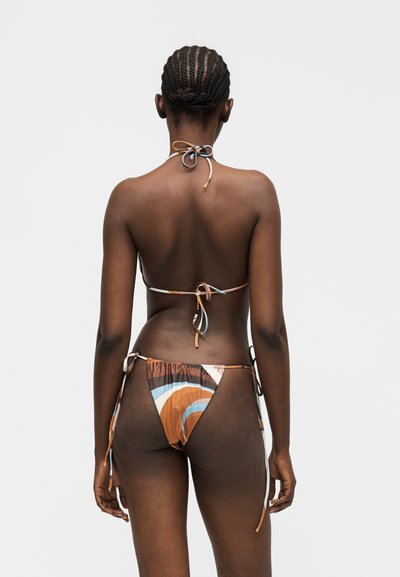 Γυναίκα με πλεξούδες, φορώντας πολύχρωμο μπικίνι με κορδόνια δεμένα, στέκεται με την πλάτη, σε απλό ανοιχτό γκρι φόντο.