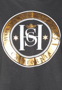 Svart bomulls T-shirt med en stor rund logotyp i vitt och guld, med texten "HOLLISTER SOUTHERN CALIFORNIA" runt kanten.