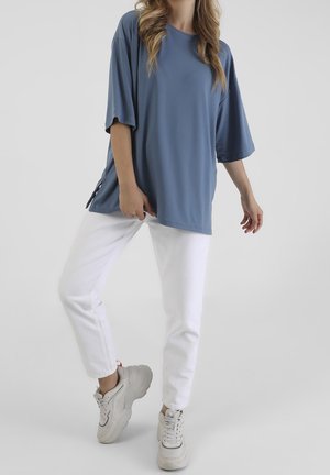 Person iført løs blå kortærmet skjorte, hvide bukser og hvide chunky sneakers, poserer mod en ensfarvet lys baggrund.