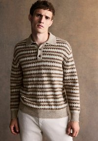 Strickpullover mit einem beigen und braunen geometrischen Muster, mit Kragen und drei Knöpfen an der Vorderseite, kombiniert mit hellen Hosen.