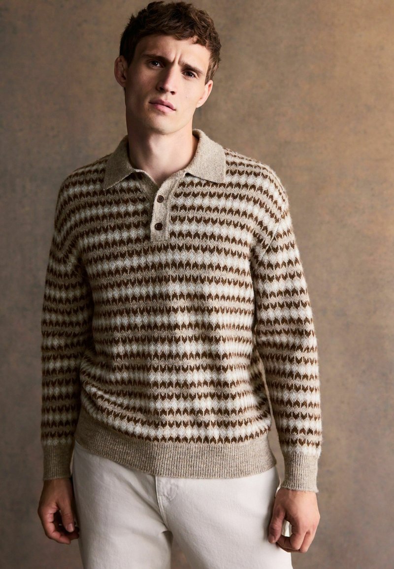 Strickpullover mit einem beigen und braunen geometrischen Muster, mit Kragen und drei Knöpfen an der Vorderseite, kombiniert mit hellen Hosen.